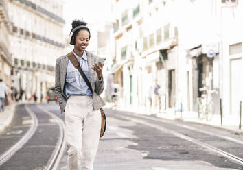 Lächelnde junge Frau mit Kopfhörern und Mobiltelefon in der Stadt unterwegs, Lissabon, Portugal - UUF19230