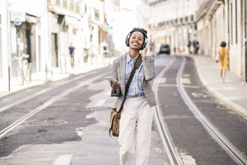 Glückliche junge Frau mit Kopfhörern und Mobiltelefon in der Stadt unterwegs, Lissabon, Portugal - UUF19232