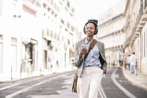 Lächelnde junge Frau mit Kopfhörern und Mobiltelefon in der Stadt unterwegs, Lissabon, Portugal - UUF19234