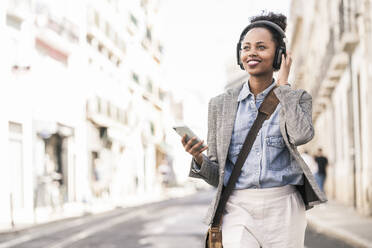 Smiling young woman with headphones and mobile phone in the city on the go, Lisbon, Portugal - UUF19235