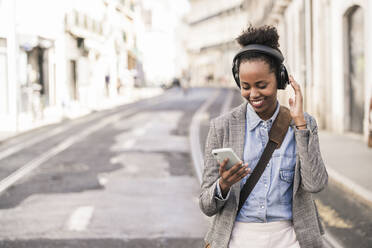 Smiling young woman with headphones and mobile phone in the city on the go, Lisbon, Portugal - UUF19237