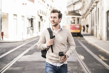 Smiling young man with backpack and coffee mug in the city on the go, Lisbon, Portugal - UUF19238