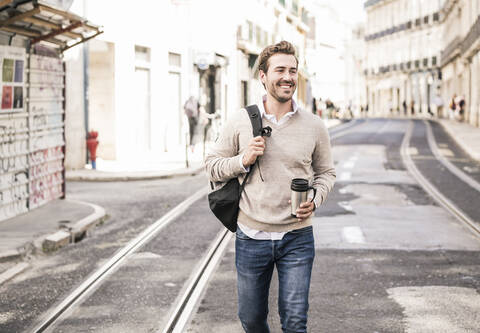 Lächelnder junger Mann mit Rucksack und Kaffeebecher in der Stadt unterwegs, Lissabon, Portugal, lizenzfreies Stockfoto
