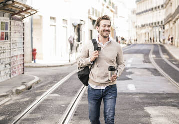 Smiling young man with backpack and coffee mug in the city on the go, Lisbon, Portugal - UUF19239