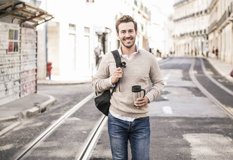 Porträt eines lächelnden jungen Mannes mit Rucksack und Kaffeebecher in der Stadt unterwegs, Lissabon, Portugal - UUF19240
