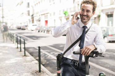 Smiling young businessman with e-scooter on the phone in the city, Lisbon, Portugal - UUF19259