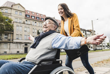 Lachende junge Frau, die einen glücklichen älteren Mann im Rollstuhl schiebt - UUF19275