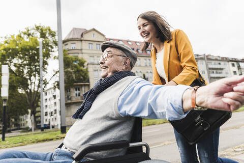 Lachende junge Frau, die einen glücklichen älteren Mann im Rollstuhl schiebt, lizenzfreies Stockfoto
