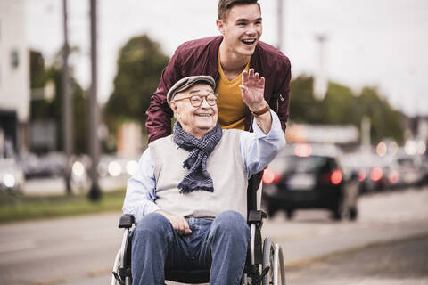 Porträt eines lachenden jungen Mannes, der einen glücklichen älteren Mann im Rollstuhl schiebt, lizenzfreies Stockfoto