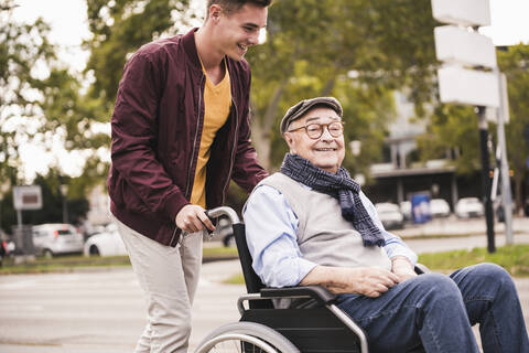 Junger Mann schiebt glücklichen älteren Mann im Rollstuhl, lizenzfreies Stockfoto