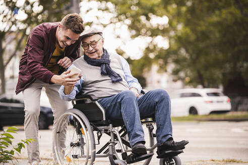 Senior man in wheelchair and his adult grandson looking together at smartphone - UUF19290