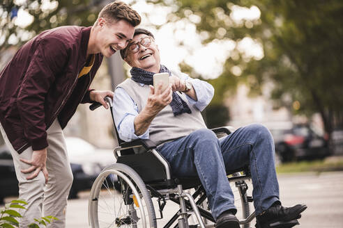 Lachender älterer Mann im Rollstuhl und sein erwachsener Enkel schauen gemeinsam auf ein Smartphone und haben Spaß - UUF19291