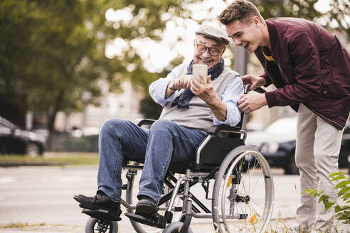 Lachender älterer Mann im Rollstuhl und sein erwachsener Enkel schauen gemeinsam auf ein Smartphone und haben Spaß - UUF19292