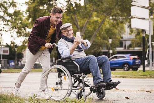 Smiling young man pushing happy senior man with smartphone in wheelchair - UUF19293