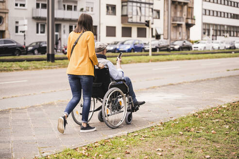 Back view of young woman pushing senior man with smartphone in wheelchair on pavement - UUF19296