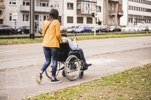 Rückenansicht einer jungen Frau, die einen älteren Mann mit Smartphone im Rollstuhl auf dem Bürgersteig schiebt - UUF19296