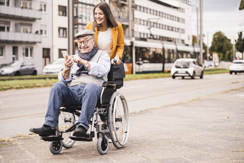 Smiling young woman pushing happy senior man with smartphone in wheelchair - UUF19297