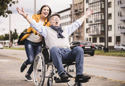 Portrait of young woman pushing senior man in wheelchair on pavement - UUF19300