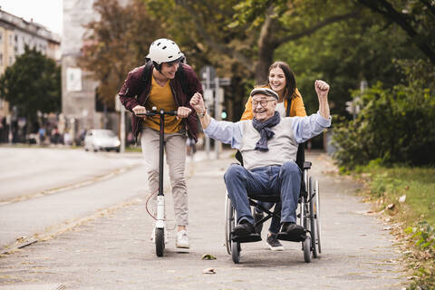 Älterer Mann im Rollstuhl vergnügt sich mit seinen erwachsenen Enkelkindern im Freien, lizenzfreies Stockfoto