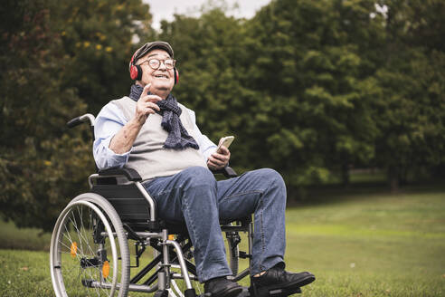 Porträt eines glücklichen älteren Mannes, der im Rollstuhl sitzt und mit Smartphone und Kopfhörern Musik hört - UUF19309