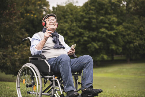 Portrait of happy senior man sitting in wheelchair listening music with smartphone and headphones - UUF19309