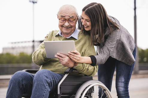 Senior man sitting in wheelchair and adult granddaughter looking together at digital tablet - UUF19313