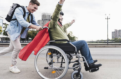 Young man pushing senior man sitting in a wheelchair dressed up as superhero - UUF19315