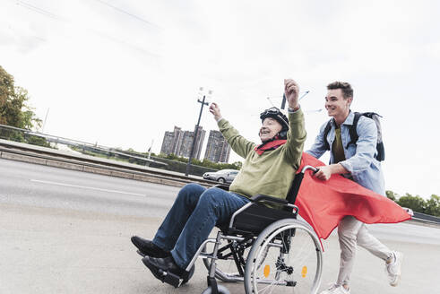 Young man pushing senior man sitting in a wheelchair dressed up as superhero - UUF19316