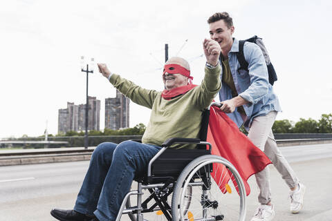 Junger Mann, der einen älteren Mann im Rollstuhl schiebt, der als Superheld verkleidet ist, lizenzfreies Stockfoto