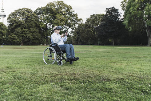 Senior man sitting in wheelchairin a park using Virtual Reality Glasses - UUF19350