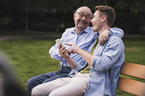 Älterer Mann und Enkel mit Smartphone sitzen zusammen auf einer Parkbank und haben Spaß, lizenzfreies Stockfoto