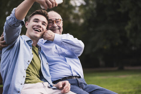 Älterer Mann und Enkel sitzen zusammen auf einer Parkbank und machen ein Selfie mit Smartphone, lizenzfreies Stockfoto