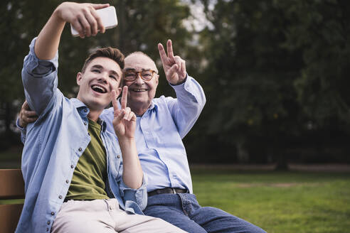 Senior man and grandson sitting together on a park bench taking selfie with smartphone - UUF19358