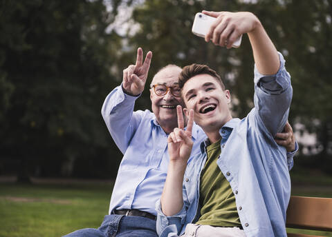 Senior man and grandson sitting together on a park bench taking selfie with smartphone stock photo