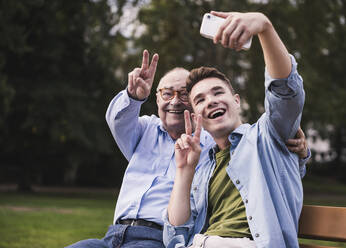 Älterer Mann und Enkel sitzen zusammen auf einer Parkbank und machen ein Selfie mit Smartphone - UUF19359