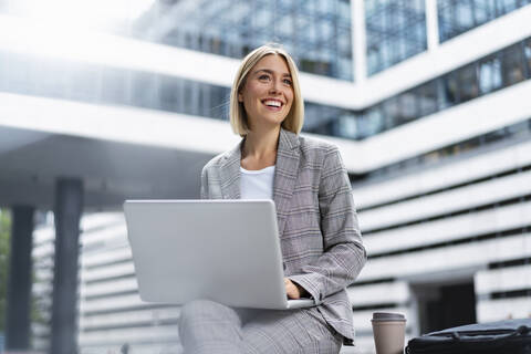 Smiling young businesswoman using laptop in the city stock photo