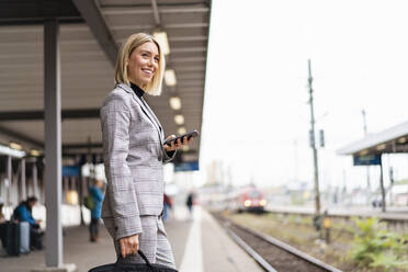 Lächelnde junge Geschäftsfrau mit Mobiltelefon am Bahnhof - DIGF08661