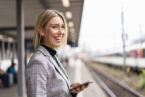 Glückliche junge Geschäftsfrau mit Mobiltelefon am Bahnhof - DIGF08663