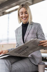 Lächelnde junge Geschäftsfrau beim Zeitungslesen am Bahnhof - DIGF08664