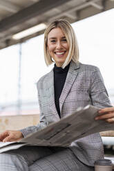 Porträt einer lächelnden jungen Geschäftsfrau mit Zeitung auf dem Bahnhof - DIGF08665