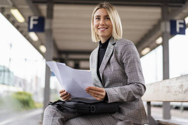 Lächelnde junge Geschäftsfrau mit Papieren auf dem Bahnhof - DIGF08667