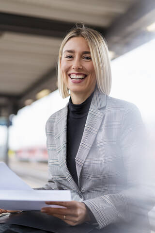 Porträt einer glücklichen jungen Geschäftsfrau mit Papieren auf dem Bahnhof, lizenzfreies Stockfoto