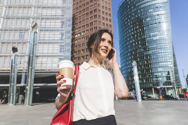 Lächelnde junge Geschäftsfrau mit Kaffee zum Mitnehmen beim Telefonieren in der Stadt, Berlin, Deutschland - WPEF02185