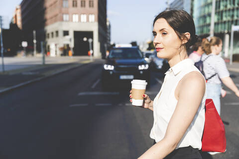 Junge Geschäftsfrau mit Kaffee zum Mitnehmen beim Überqueren der Straße in der Stadt, Berlin, Deutschland, lizenzfreies Stockfoto