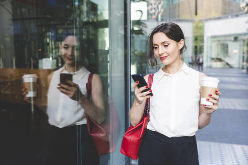 Junge Geschäftsfrau mit Kaffee zum Mitnehmen und Mobiltelefon, das sich in einer Glasfront spiegelt, Berlin, Deutschland - WPEF02191