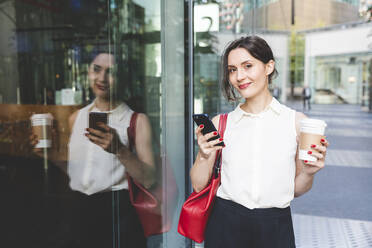 Porträt einer jungen Geschäftsfrau mit Kaffee zum Mitnehmen und Mobiltelefon, das sich in einer Glasfront spiegelt, Berlin, Deutschland - WPEF02192