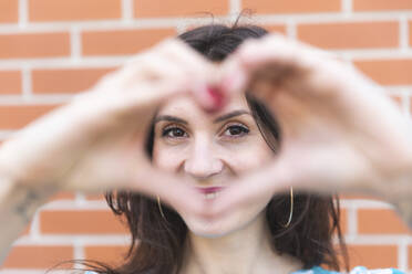 Portrait of smiling young woman looking at camera through heart-shaped hands - WPEF02199