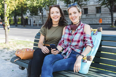 Porträt von zwei jungen Frauen auf einer Bank sitzend mit Skateboard und Mobiltelefon - WPEF02203