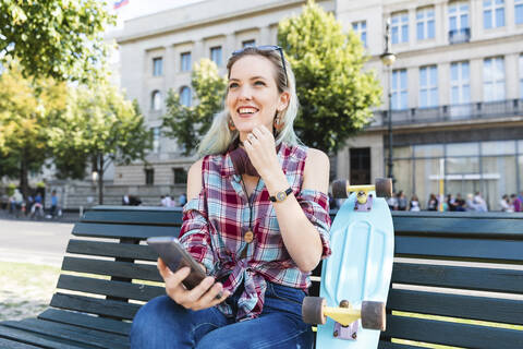 Porträt einer lächelnden jungen Frau auf einer Bank sitzend mit Skateboard und Mobiltelefon, lizenzfreies Stockfoto