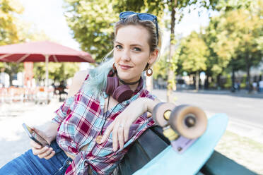 Portrait of smiling young woman sitting on bench with skateboard and smartphone - WPEF02205